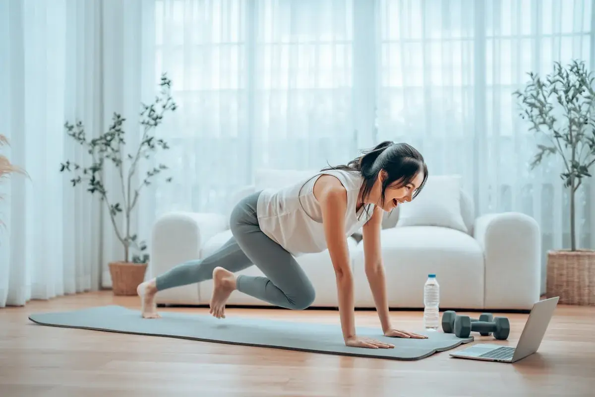 Kobieta wykonuje ćwiczenie "mountain climbers" na macie, ucząc się, trening cardio co to jest, z laptopem i hantlami obok.