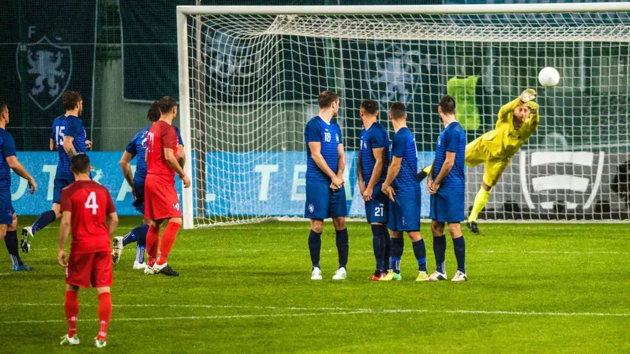 Bramkarz w żółtym stroju broni strzał. Piłkarze w niebieskich i czerwonych strojach czekają na rozwój akcji. Zastanawiasz się, ile trwa mecz piłki nożnej?