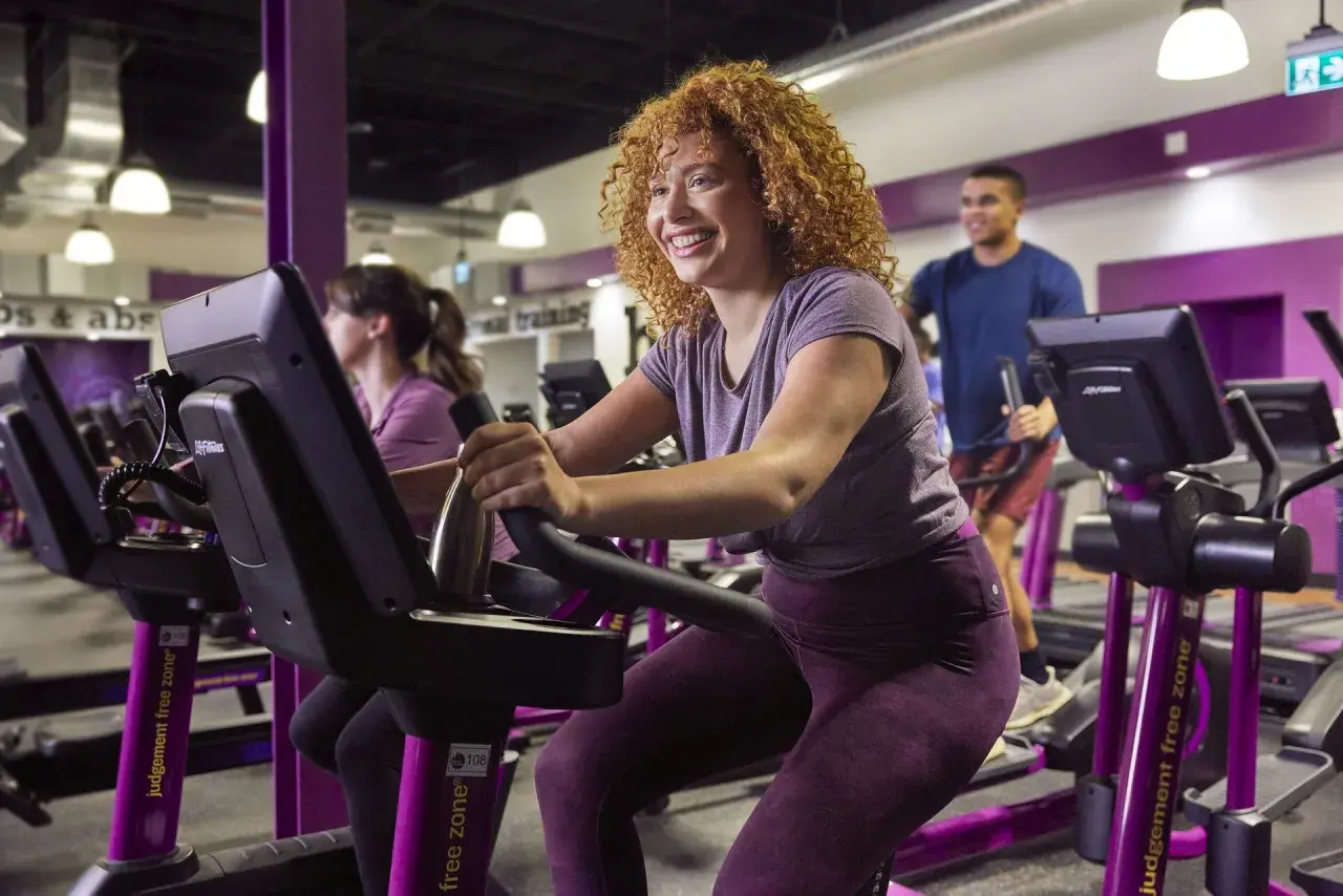 Uśmiechnięta kobieta z kręconymi włosami wykonuje trening cardio na rowerku stacjonarnym. To świetny sposób na poprawę kondycji!